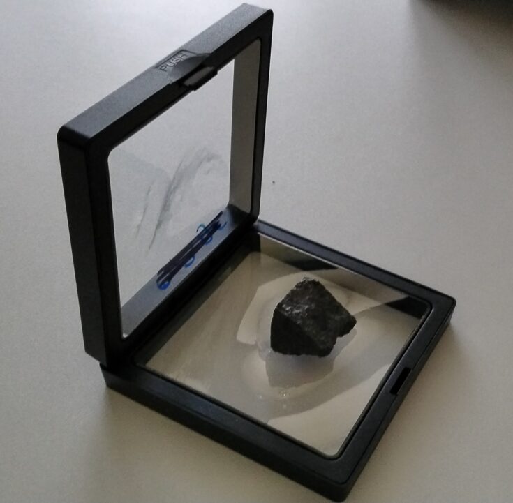 Petite roche noire dans une petite boîte noire avec vitre transparente, posé sur une table.